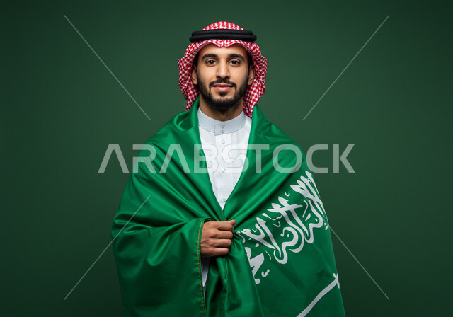 النظر الى الكاميرا بتعابير السرور والفخر بالانتماء للوطن، بورتريه رجل عربي خليجي سعودي مبتسم يرتدي الشماغ والثوب التقليدي يضع الراية على كتفه يحتفل باليوم الوطني السعودي 23 سبتمبر، يوم العلم 11 مارس، احتفالات ومناسبات وطنية في المملكة العربية السعودية، خلفية خضراء