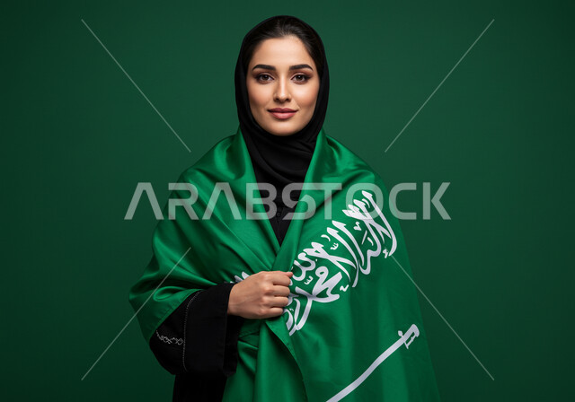 النظر الى الكاميرا بتعابير السرور والفخر بالانتماء للوطن، بورتريه إمرأة عربية خليجية سعودية مبتسمة ترتدي الحجاب والعباءة تضع الراية على كتفها تحتفل باليوم الوطني السعودي 23 سبتمبر، يوم العلم 11 مارس، احتفالات ومناسبات وطنية في المملكة العربية السعودية، خلفية خضراء