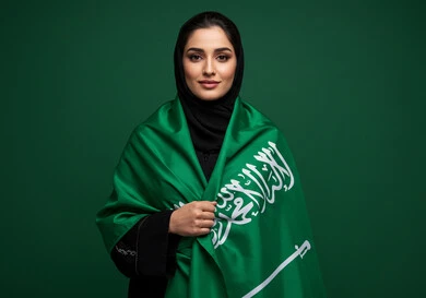 النظر الى الكاميرا بتعابير السرور والفخر بالانتماء للوطن، بورتريه إمرأة عربية خليجية سعودية مبتسمة ترتدي الحجاب والعباءة تضع الراية على كتفها تحتفل باليوم الوطني السعودي 23 سبتمبر، يوم العلم 11 مارس، احتفالات ومناسبات وطنية في المملكة العربية السعودية، خلفية خضراء