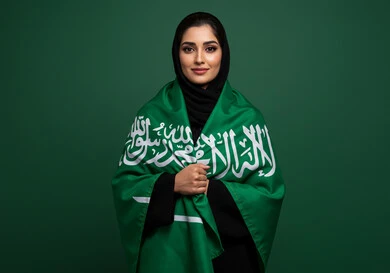 النظر الى الكاميرا بتعابير السرور والفخر بالانتماء للوطن، بورتريه إمرأة عربية خليجية سعودية مبتسمة ترتدي الحجاب والعباءة تضع الراية على كتفها تحتفل باليوم الوطني السعودي 23 سبتمبر، يوم العلم 11 مارس، احتفالات ومناسبات وطنية في المملكة العربية السعودية، خلفية خضراء