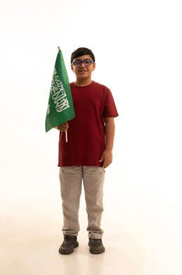 الاحتفالات بالمناسبات والأعياد الشعبية الوطنية، إحياء ذكرى يوم العلم السعودي، تربية الأبناء على حب الوطن، بورتريه لولد عربي خليجي سعودي يرتدي الزي الكاجوال الاحتفال باليوم الوطني السعودي ويحمل راية المملكة العربية السعودية، الجسم بالطول الكامل، خلفية بيضاء