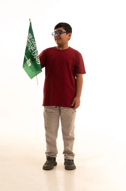Raising children to love their country, celebrating national holidays and popular occasions, commemorating Saudi Arabia's Flag Day, a portrait of an Arab Gulf Saudi boy wearing casual attire celebrating Saudi National Day and holding the flag of the Kingdom of Saudi Arabia, full-body, white background.