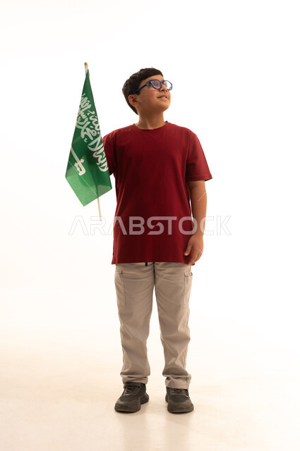 Commemorating Saudi Flag Day, celebrations of national popular occasions and holidays, raising children to love the homeland, a portrait of an Arab Gulf Saudi boy wearing casual attire celebrating Saudi National Day and holding the flag of the Kingdom of Saudi Arabia, full-body, white background.