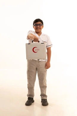 شكل حقيبة الاسعافات الاولية، النظر الى الكاميرا بتعابير الشغف لتحقيق حلم التمريض، تقديم الرعاية والخدمات الصحية، بورتريه لفتى عربي خليجي سعودي مبتسم يرتدي المعطف الأبيض يقف ويحمل حقيبة الاسعافات الأولية، اختيار وتحديد المهنة المستقبلية منذ الصغر، خلفية بيضاء