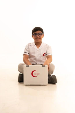 النظر الى الكاميرا بتعابير الشغف لتحقيق حلم التمريض، تقديم الرعاية والخدمات الصحية، بورتريه لفتى عربي خليجي سعودي مبتسم يرتدي المعطف الأبيض يجلس على الارض يتفقد محتويات حقيبة الاسعافات الأولية، اختيار وتحديد المهنة المستقبلية منذ الصغر، خلفية بيضاء