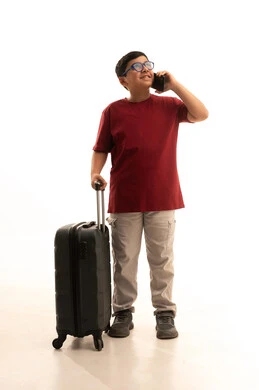 Talking with friends and family before traveling, using technologies and travel applications and hotel bookings, the concept of tourism and travel, a portrait of an Arab Gulf Saudi boy wearing casual attire, red pants and a t-shirt, holding a mobile phone and a travel bag, white background.
