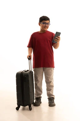 The use of tourism technologies and hotel booking applications, the concept of tourism and travel, a portrait of a young Arab Gulf Saudi boy wearing casual attire, red pants and a t-shirt, holding a mobile phone and a travel bag, talking to friends via social media, white background.