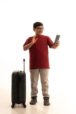 The concept of tourism and travel, a portrait of a young Arab Gulf Saudi boy wearing casual attire, pants and a red t-shirt, holding a mobile phone and a travel bag, using tourism technologies and applications and hotel bookings, talking to friends via social media, white background.