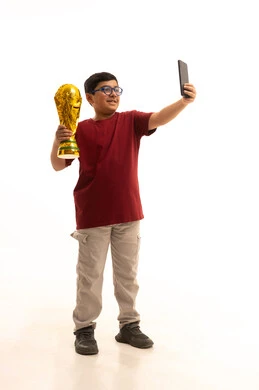 التقاط سيلفي مع الكأس الذهبي، مفهوم الربح والفوز، ايماءات السعادة والفرح, بورتريه لولد عربي خليجي سعودي يرتدي الزي الكاجوال سروال وفانيلا حمراء يضع على رأسه كأس العالم لرياضة كرة القدم، نشاط بدني للمحافظة على صحة الاجسام و اللياقة، الجسم بالطول الكامل، خلفية بيضاء