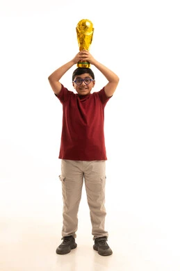 مفهوم الربح والفوز، ايماءات السعادة والفرح, بورتريه لولد عربي خليجي سعودي يرتدي الزي الكاجوال سروال وفانيلا حمراء يضع على رأسه كأس العالم الذهبي لرياضة كرة القدم، نشاط بدني للمحافظة على صحة الاجسام و اللياقة، الجسم بالطول الكامل، خلفية بيضاء