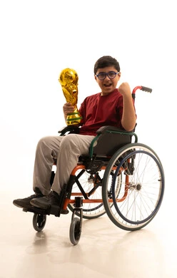 تعابير الفرح والسرور، تنمية الهوايات الرياضية لذوي الإعاقة, بورتريه لولد عربي خليجي سعودي مبتسم مقعد يرتدي الزي الكاجوال والنظارة الطبية يجلس على كرسي متحرك يحمل كأس ذهبي يرفع يده بايماءات الفوز والنجاح، نشاطات ترفيهية لذوي الهمم، خلفية بيضاء