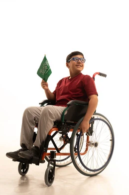 Participation of people with determination in holidays and national events, commemorating the Saudi National Day on September 23, a portrait of an Arab Gulf Saudi boy wearing casual attire and glasses sitting in a wheelchair holding the Saudi flag with respect and pride, expressing love for the homeland and belonging to it, white background.