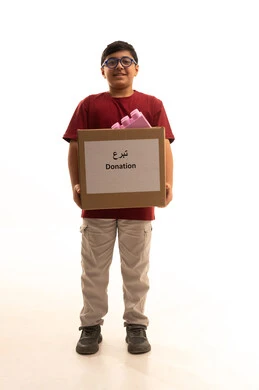 توفير وسائل التسلية والترفيه، التبرع بألعاب أطفال لادخال البهجة على قلب الصغار،المشاركة في الثواب، المساعدة على توفير الاحتياجات، بورتريه لولد عربي خليجي سعودي يرتدي الزي الكاجوال ويضع نظارات طبية يقوم بحمل صندوق التبرع، الجسم بالطول الكامل، خلفية بيضاء