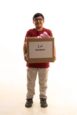 التبرع بألعاب أطفال لادخال البهجة على قلب الصغار، توفير وسائل التسلية والترفيه، المشاركة في الثواب، المساعدة على توفير الاحتياجات، بورتريه لولد عربي خليجي سعودي يرتدي الزي الكاجوال ويضع نظارات طبية يقوم بحمل صندوق التبرع، الجسم بالطول الكامل، خلفية بيضاء