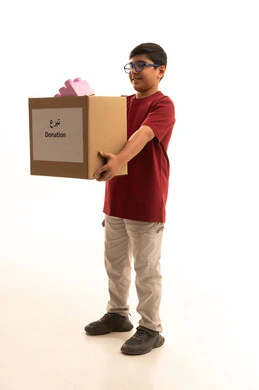 Providing means of entertainment and amusement, donating children's toys to bring joy to the hearts of the little ones, participating in rewards, helping to provide needs, a portrait of an Arab Gulf Saudi boy wearing casual attire and wearing prescription glasses carrying a donation box, full-body, white background.