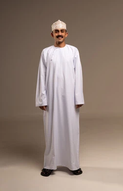 الوقوف باستقامة والنظر للكاميرا بإيمات السرور والسعادة، بورتريه رجل عربي خليجي عماني يرتدي الدشداشة والكمة يقف ويبتسم، مفهوم الهيبة والاناقة، تمثيل الرجولة العربية الأصيلة، المحافظة على اللباس التقليدي، الجسم بالطول الكامل، خلفية بيج