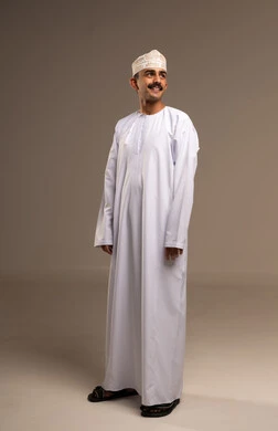 مفهوم لغة الجسد وتعابيرالوجه، تمثيل الرجولة العربية الأصيلة، الوقوف باستقامة والنظر للاعلى بإيمات السرور والسعادة، بورتريه رجل عربي خليجي عماني يرتدي الدشداشة والكمة يقف ويبتسم، مفهوم الهيبة والاناقة، المحافظة على اللباس التقليدي، الجسم بالطول الكامل، خلفية بيج