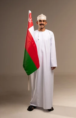 الاحتفال بذكرى اليوم الوطني العماني، المشاركة بالاعياد والمناسبات الوطنية، الاعتزاز والفخر بالرموز والراية العمانية، بورتريه لشاب عربي خليجي عماني يرتدي الدشداشة والكمه يحمل بيده علم سلطنة عمان، مفهوم حب الوطن والإنتماء إليه، الجسم بالطول الكامل، خلفية بيج