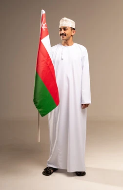 The concept of love for the homeland and belonging to it, celebrating the anniversary of Oman's National Day, participating in national holidays and events, pride and honor in the symbols and flag of Oman, a portrait of a young Arab Gulf Omani man wearing a dishdasha and kumma holding the flag of the Sultanate of Oman in his hand, full-body, beige background.