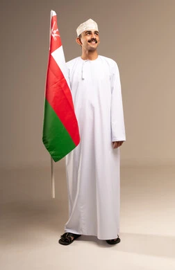المشاركة بالاعياد والمناسبات الوطنية، الاحتفال بذكرى اليوم الوطني العماني، الاعتزاز والفخر بالرموز والراية العمانية، بورتريه لشاب عربي خليجي عماني يرتدي الدشداشة والكمه يحمل بيده علم سلطنة عمان، مفهوم حب الوطن والإنتماء إليه، الجسم بالطول الكامل، خلفية بيج