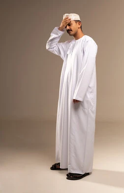 وضع اليد على الرأس بسبب الألم، التعب وتحمل الالام ، الذهاب للطبيب لمعالجة الاوجاع، تمثيل الرجولة العربية،  بورتريه  لرجل عربي خليجي عماني يرتدي الدشداشة والكمة، مفهوم الاهتمام بالاناقة والمظهر الخارجي، المحافظة على لبس اللباس التقليدي، الجسم بالطول الكامل، خلفية بيج