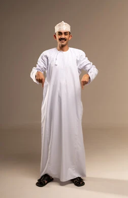 Pointing to the feet, using the index finger in movements and expressions, expressions of joy and happiness, identifying a specific location or goal, the concept of elegance and masculinity, a portrait of an Omani Gulf Arab man wearing a dishdasha and kumma standing, attention to external appearance, full-body, beige background.