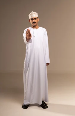 Calmness and stillness and no disturbance, the command to stop doing something, expressions of rejection and refusal, silence and not speaking, a portrait of a young Arab Gulf Omani man wearing a dishdasha and kumma raising his hand with gestures of prohibition, feelings of annoyance and anger, full-body image, beige background.