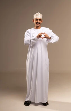 Hand gestures in the shape of a heart, expressing feelings of love and passion, the concept of affection and tenderness, a portrait of a young Arab Gulf Omani man wearing a dishdasha and kumma looking at the camera with expressions of joy and happiness, full-body image, beige background.