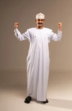 الشعور بالقوة والسرور، مفهوم اللياقة البدنية وكمال الأجسام، بناء جسم رياضي، بورتريه لشاب عربي خليجي عماني يرتدي الدشداشة والعمامة يرفع يده للاعلى يقوم بالنظر الى عضلاته، مفهوم الاهتمام بالمطهر الخارجي والاناقة، صورة الجسم بالطول الكامل، خلفية بيج