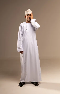 الالتهابات و المشاكل العينية و الحالات المرضية، ضرورة زيارة الطبيب والقيام بالفحوصات، الحاجة للفحص والعلاج، دخول شيء مزعج و مؤلم في العين, بورتريه لشاب عربي خليجي عماني يرتدي الدشداشة والكمه يضع يده على عينه بتعابير الانزعاج و الالم, الجسم بالطول الكامل, خلفية بيج