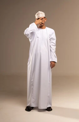 حركات اليدين والوجه تعبر عن الخجل، إيماءات تذكر شيء ما بشكل مفاجئ، بورتريه مقرب لرجل عربي خليجي عماني يرتدي الدشداشة والكمه العمانية التقليدية يقف بشكل مستقيم ويغطي عينيه بيده، الاهتمام بالمظهر الخارجي، حركات تدل على اللعب و المرح, خلفية بيج