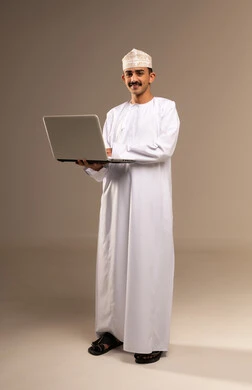 انجاز المهام والعمل عن بعد، مفهوم استخدام التقنيات الحديثة والأجهزة المتطورة ، بورتريه بالطول الكامل لشاب عربي خليجي عماني يرتدي الدشداشة والكمه يحمل بيده جهاز الحاسب المحمول تظهر عليه علامات الفرح والسرور،  مهن ووظائف مكتبيه عمانية، خلفية بيج