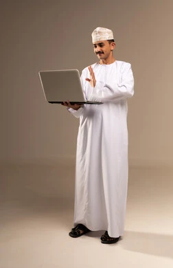 A video call on a laptop, the concept of using modern technologies and advanced devices, a full-length portrait of a young Arab Gulf Omani man wearing a dishdasha and kumma holding a laptop in his hand showing signs of joy and happiness, Omani office professions and jobs, beige background.