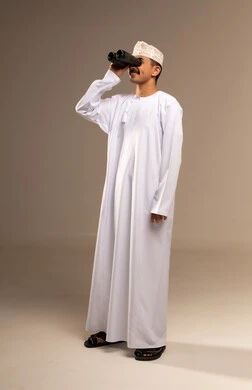 Love of exploration and adventure, clarifying vision through a telescope, a portrait of an Arab Gulf Omani man wearing a dishdasha and kumma holding a binocular in his hand and looking at something, zooming in on distant objects and places, gestures of enjoyment and pleasure, full-body image, beige background.