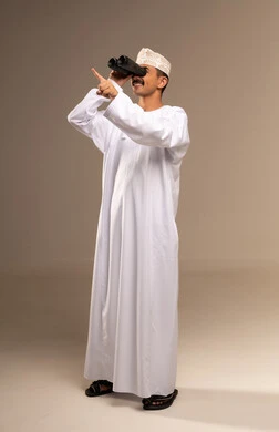 إيماءات الاستمتاع والسرور, حب الاستكشاف و المغامرة, توضيح الرؤية عبر التلسكوب, بورتريه لرجل عربي خليجي عماني يرتدي الدشداشة والكمه يحمل بيده منظار ثنائي العينين و ينظر الى شيء ما, تقريب الأشياء والأماكن البعيدة، صورة الجسم بالطول الكامل, خلفية بيج