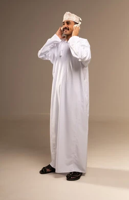 الاستمتاع بسماع الموسيقى المفضلة, استخدام التقنيات الحديثة للتسلية والترفيه, قضاء الوقت الممتع, الاندماج مع الألحان الموسيقية, بورتريه لشاب عربي خليجي عماني مبتسم يرتدي الدشداشة والكمة يضع سماعات الرأس الصوتية ويرفع يده للأعلى, الجسم بالطول الكامل, خلفية بيج