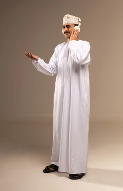 استخدام التقنيات الحديثة للتسلية والترفيه, الاستمتاع بسماع الموسيقى المفضلة,  قضاء الوقت الممتع, الاندماج مع الألحان الموسيقية, بورتريه لشاب عربي خليجي عماني مبتسم يرتدي الدشداشة والكمة يضع سماعات الرأس الصوتية ويرفع يده للأعلى, الجسم بالطول الكامل, خلفية بيج