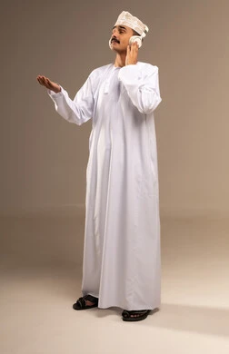 Spending enjoyable time, using modern technologies for entertainment and amusement, enjoying listening to favorite music, immersing in musical melodies, a portrait of a smiling young Arab Gulf Omani man wearing a dishdasha and kumma, wearing headphones and raising his hand up, full body, beige background.