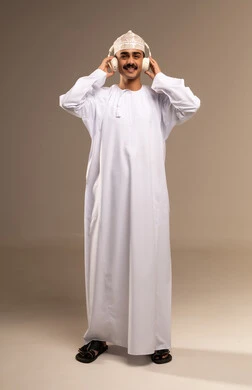الاندماج مع الألحان الموسيقية, قضاء الوقت الممتع, استخدام التقنيات الحديثة للتسلية والترفيه, الاستمتاع بسماع الموسيقى المفضلة,   بورتريه لشاب عربي خليجي عماني مبتسم يرتدي الدشداشة والكمة يضع سماعات الرأس الصوتية ويرفع يده للأعلى, الجسم بالطول الكامل, خلفية بيج