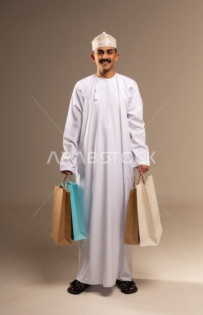 الاستفادة من العروض و التخفيضات الموسمية في الاسواق, بورتريه لشاب عربي خليجي عماني مبتسم يرتدي الدشداشة والكمه يحمل بيده اكياس التسوق الملونة ينظر الى الكاميرا بايماءات السرور, متعة في التسويق و اقتناء المستلزمات و الهدايا, الجسم بالطول الكامل, خلفيه بيج