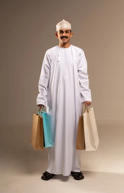 الاستفادة من العروض و التخفيضات الموسمية في الاسواق, بورتريه لشاب عربي خليجي عماني مبتسم يرتدي الدشداشة والكمه يحمل بيده اكياس التسوق الملونة ينظر الى الكاميرا بايماءات السرور, متعة في التسويق و اقتناء المستلزمات و الهدايا, الجسم بالطول الكامل, خلفيه بيج