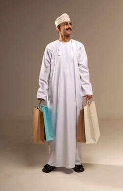 متعة في التسويق و اقتناء المستلزمات و الهدايا, الاستفادة من العروض و التخفيضات الموسمية في الاسواق, بورتريه لشاب عربي خليجي عماني مبتسم يرتدي الدشداشة والكمه يحمل بيده اكياس التسوق الملونة ينظر الى الكاميرا بايماءات السرور,  الجسم بالطول الكامل, خلفيه بيج
