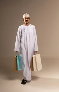تعابير وحركات الحماس للتسوق،  شراء المنتجات و الدفع عن بعد, الاستفادة من العروض و التخفيضات الموسمية في الاسواق, بورتريه لشاب عربي خليجي عماني مبتسم يرتدي الدشداشة والكمه يحمل بيده اكياس التسوق الملونة ينظر الى الكاميرا بايماءات السرور,  الجسم بالطول الكامل, خلفيه بيج