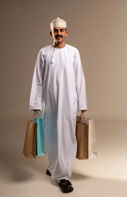 شراء الأغراض ومتعة التسوق في المولاتالاستفادة من العروض و التخفيضات الموسمية في الاسواق, بورتريه لشاب عربي خليجي عماني مبتسم يرتدي الدشداشة والكمه يحمل بيده اكياس التسوق الملونة ينظر الى الكاميرا بايماءات السرور,  الجسم بالطول الكامل, خلفيه بيج