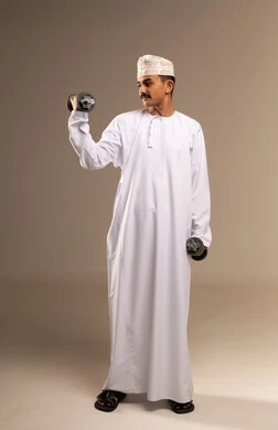 Physical activity to maintain health and physical fitness, a portrait of a young Arab Gulf Omani man wearing a dishdasha and kumma practicing exercises with dumbbells, bodybuilding and muscle building, lifting heavy weights, gestures of effort and fatigue, full-body shot, beige background.