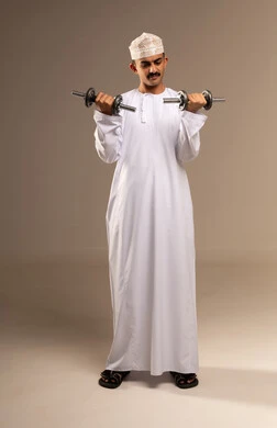 Bodybuilding and muscle building is a physical activity to maintain health and physical fitness, featuring a portrait of a young Arab Gulf Omani man wearing a dishdasha and kumma practicing exercises with dumbbells, lifting heavy weights, showing gestures of effort and fatigue, full-body shot, beige background.