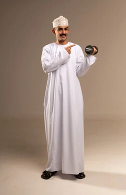 Lifting heavy weights, gestures of effort and fatigue, bodybuilding and muscle building, physical activity to maintain health and physical fitness, a portrait of a young Arab Gulf Omani man wearing a dishdasha and kumma exercising with dumbbells, full-body, beige background.