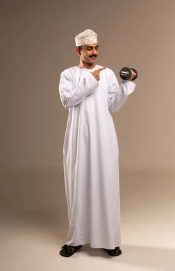 Building and strengthening body muscles, lifting heavy weights, gestures of effort and fatigue, physical activity to maintain health and physical fitness, a portrait of a young Arab Gulf Omani man wearing a dishdasha and kumma exercising with dumbbells, full-body, beige background.