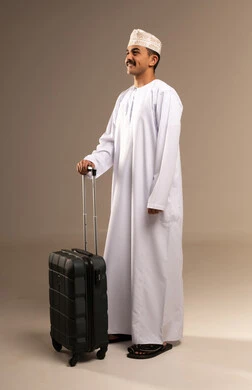 Spending enjoyable times during the vacation, the concept of tourism and travel, a portrait of a young Arab Gulf Omani man wearing a dishdasha and a kumma holding a travel bag in his hand, looking at something with expressions of excitement and joy, preparing for the time of the trip, full-body, beige background.