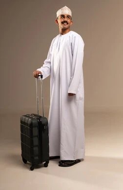 Looking at something with expressions of excitement and joy, spending enjoyable times during the vacation, the concept of tourism and travel, a portrait of a young Arab Gulf Omani man wearing a dishdasha and a kumma holding a travel bag in his hand, preparing for the time of the trip, full-body, beige background.